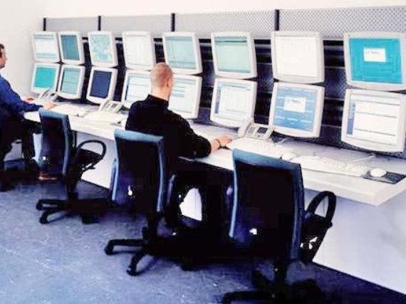Dwie osoby siedzące przy biurkach na których stoi 16 monitorów 8 klawiatur i przy biurkach stoją 4 krzesła